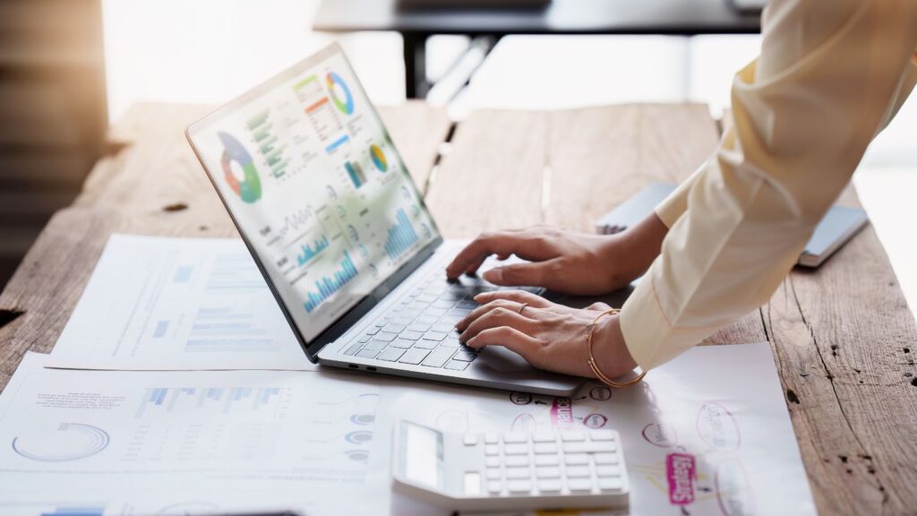 Person analyzing data on a laptop displaying colorful charts and graphs, surrounded by printed reports and a calculator on a wooden desk—representing business analytics, workflow automation, and financial reporting.