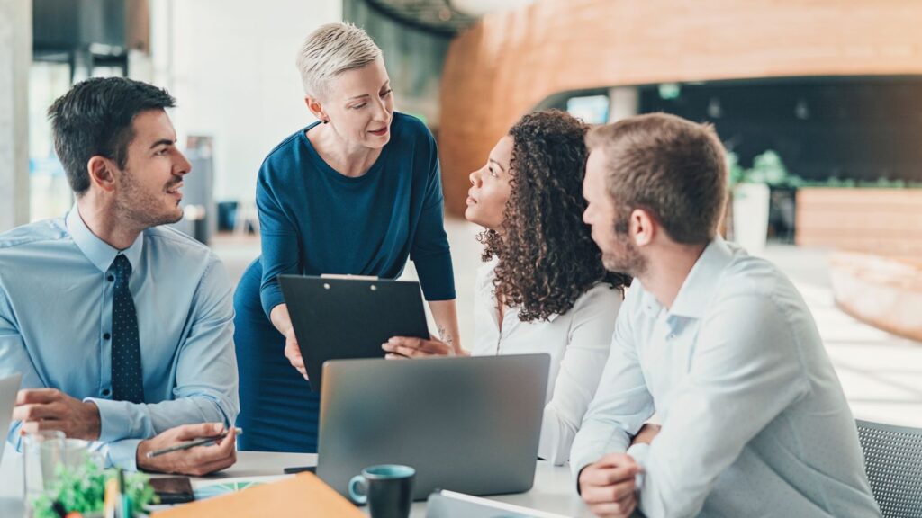 Female business leader standing with a clipboard, engaging in discussion with three colleagues around a table with laptops, representing leadership, collaboration, and decision-making in a modern workplace.
