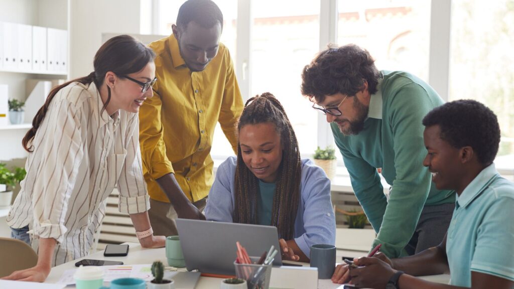 Benefits of Business Central SaaS: Enhanced Reporting, Collaboration & Continuous Security 2 Diverse business team collaborating around a laptop in a bright modern office, representing teamwork and improved collaboration—one of the key benefits of Business Central SaaS.