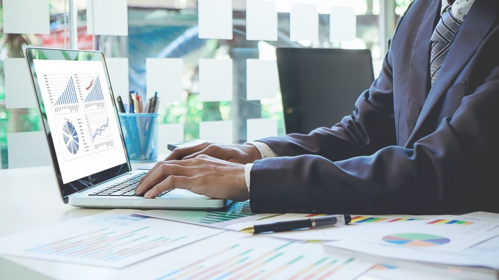 ERP Knowledge Transfer in Manufacturing: How to Prevent Disruption When People Leave 4 A business professional in a suit working on a laptop displaying charts and graphs, with printed financial reports and a pen on the desk, representing data analysis, business strategy, or financial planning.