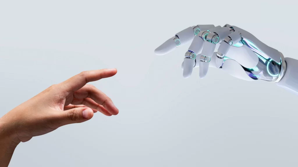 Close-up of a human hand reaching toward a white robotic hand, fingertips nearly touching against a light gray background, symbolizing human–AI collaboration.