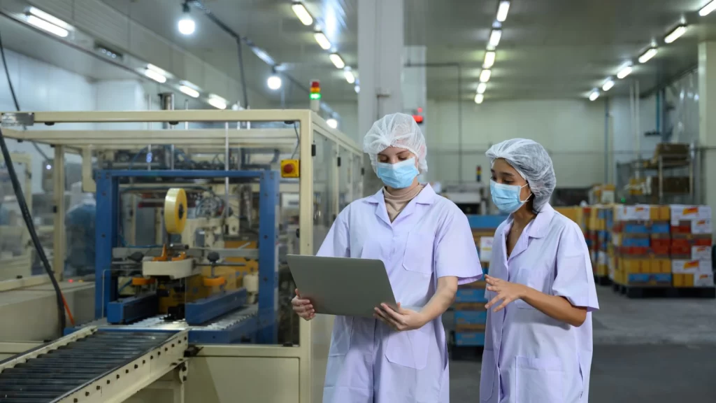 Quality management in a food manufacturing facility, with QA team members reviewing production data on a tablet while monitoring packaging operations on the factory floor.