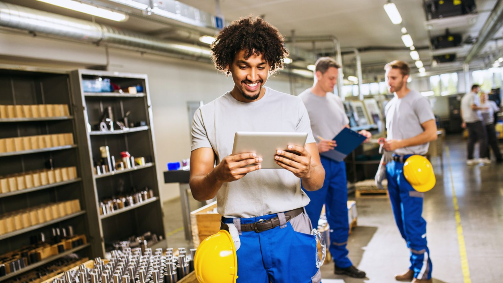 Manufacturing workers on a factory floor, with a technician using a tablet to review production data while team members coordinate tasks, illustrating workforce planning, labor tracking, and digital operations in manufacturing.